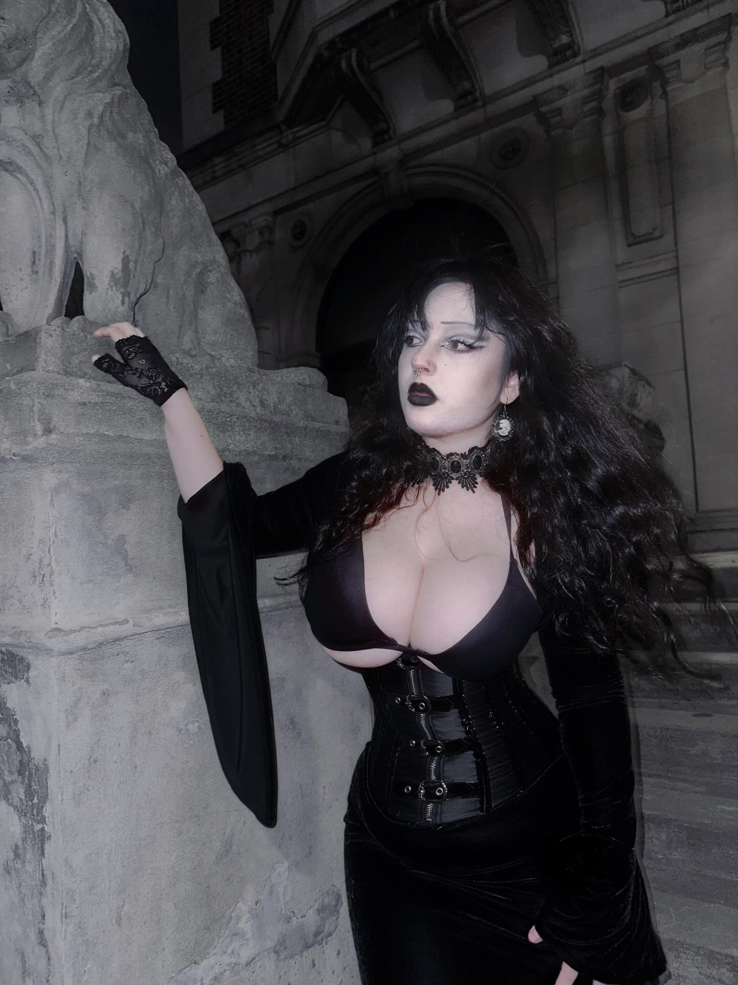 Ghoulishbats in black patent corset and lace cameo choker leaning against stone statue at night