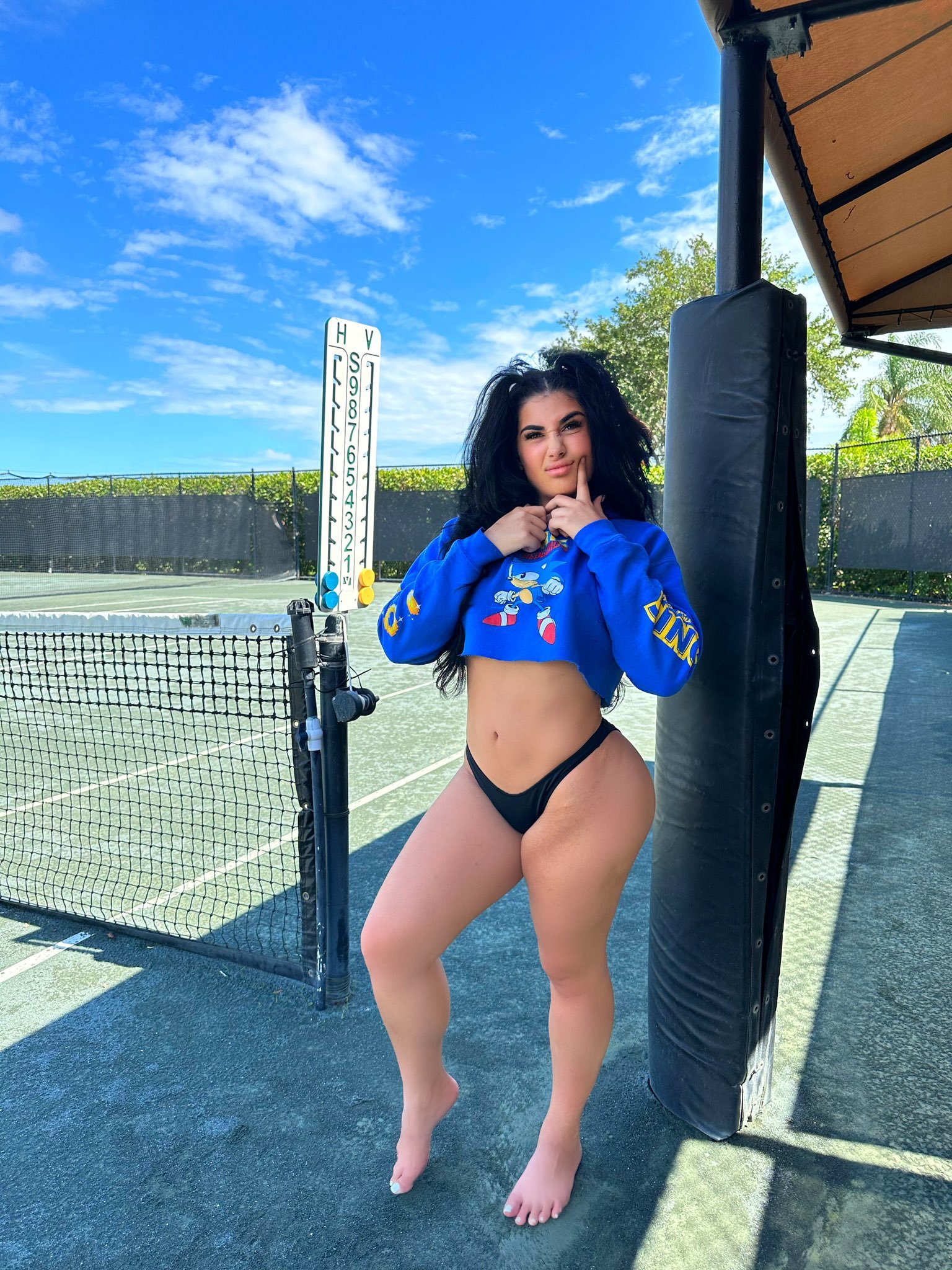Lord Len scrunches her nose and presses a finger to her cheek while posing by a tennis net in a cropped blue Sonic the Hedgehog hoodie and black thong
